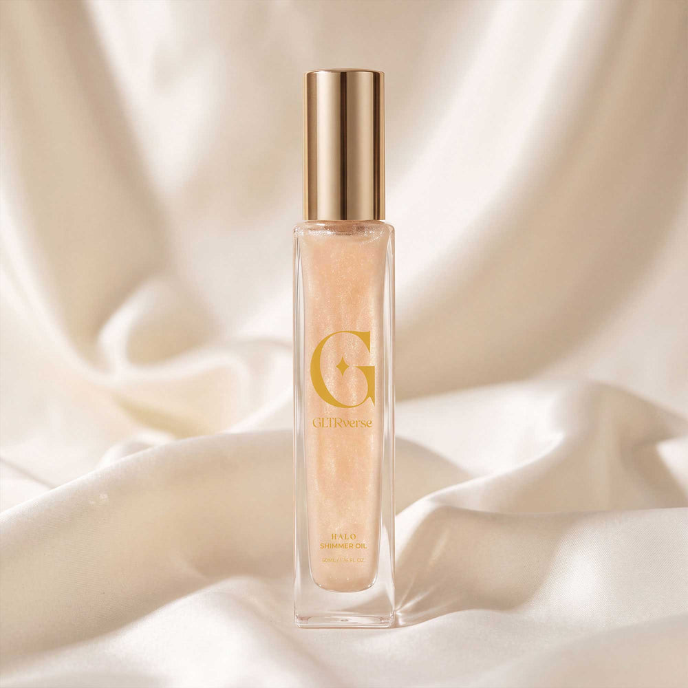 GLTRverse Halo warm gold shimmer body oil in a tall slim clear glass bottle with a gold pump cap, standing on cream silk