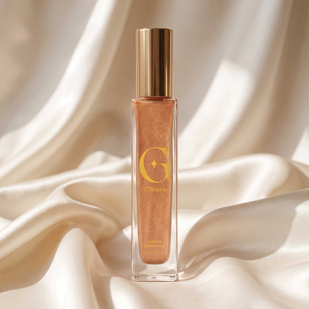 GLTRverse Amber bronze-gold shimmer body oil in a tall slim clear glass bottle with a gold pump cap, on cream silk
